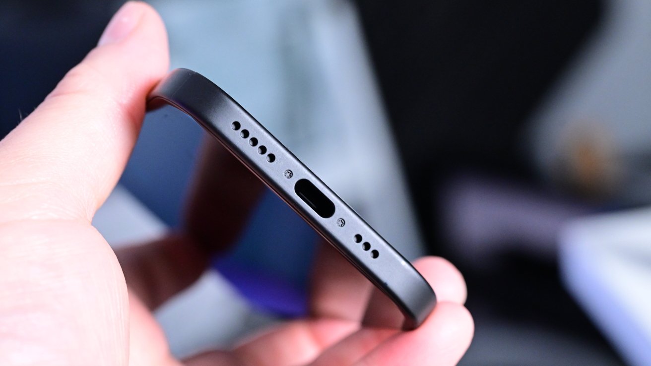 A hand holds a smartphone focusing on its bottom edge, showing a USB-C charging port and speaker holes.