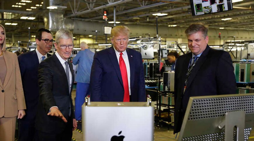 Group of people in a factory setting looking at a high-tech machine with an Apple logo. Factory equipment and industrial ceiling visible in the background.