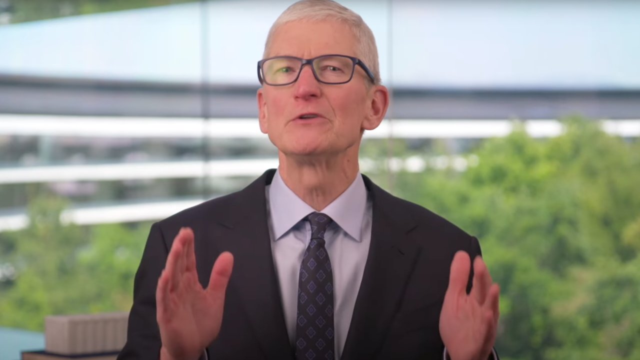 Man in a suit gestures while speaking, set against a blurred backdrop of greenery and a modern building with large windows.