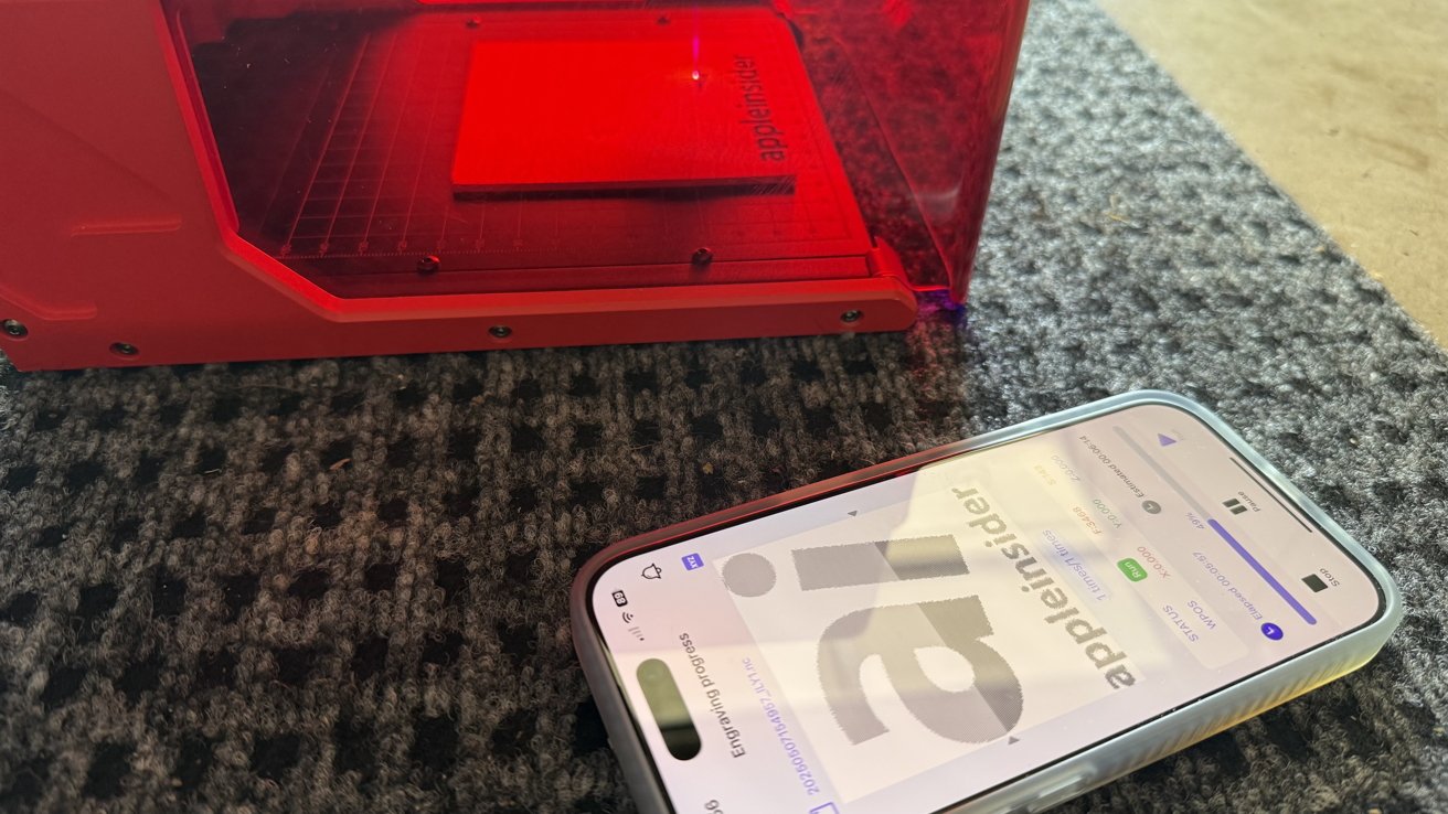 A red 3D printer with a phone displaying progress on a carpeted floor.