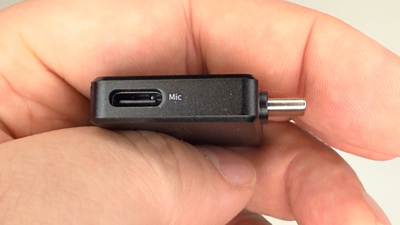 Hand holding a small black electronic device with a microphone slot and charging pin visible.