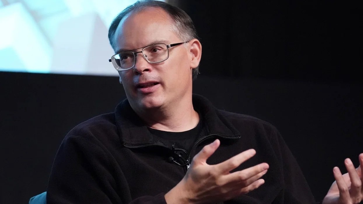 A person in glasses and a dark jacket gestures while speaking, seated against a dark background.