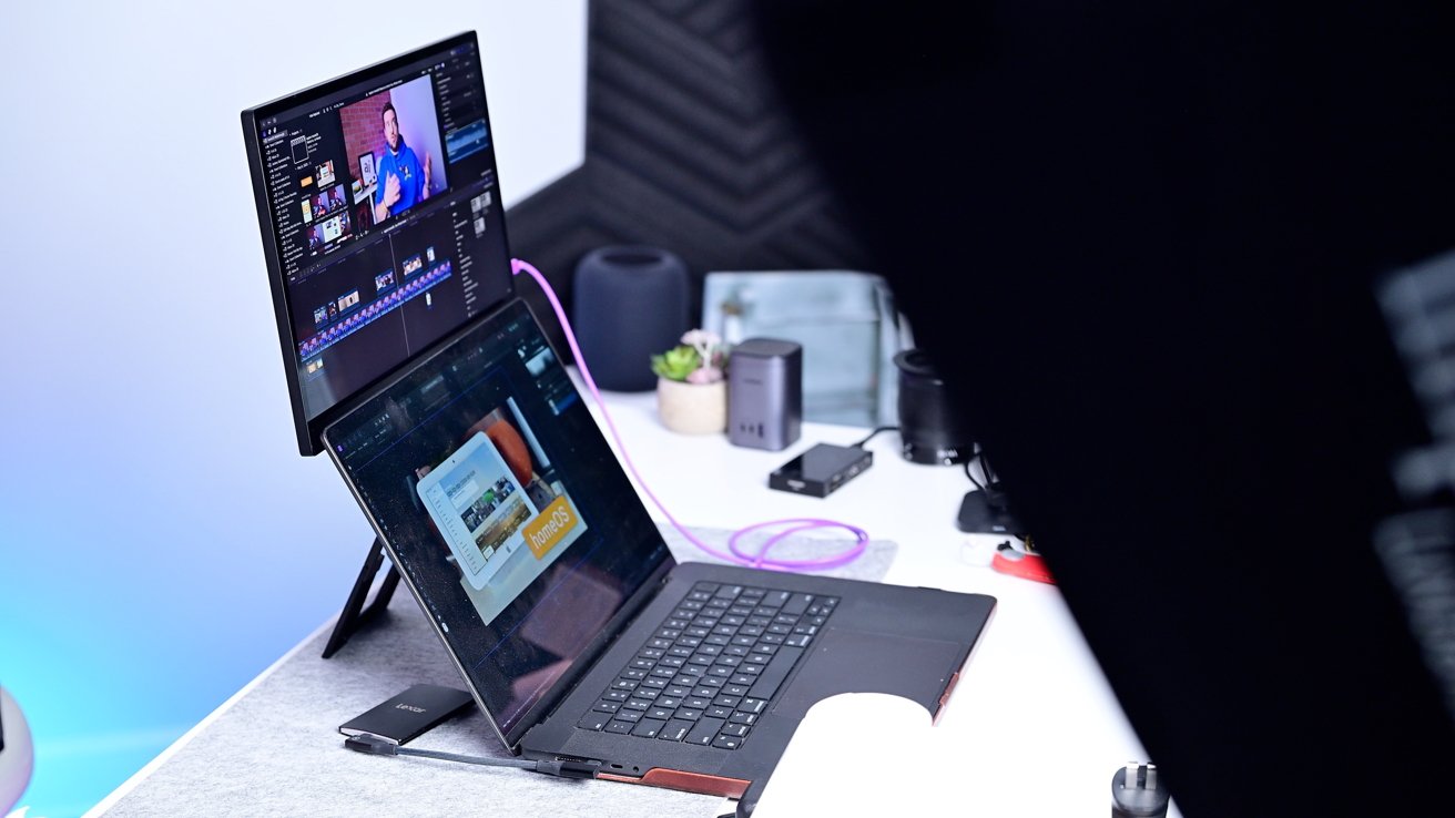 Laptop and monitor on a desk showing video editing software, surrounded by various tech accessories and a small plant.
