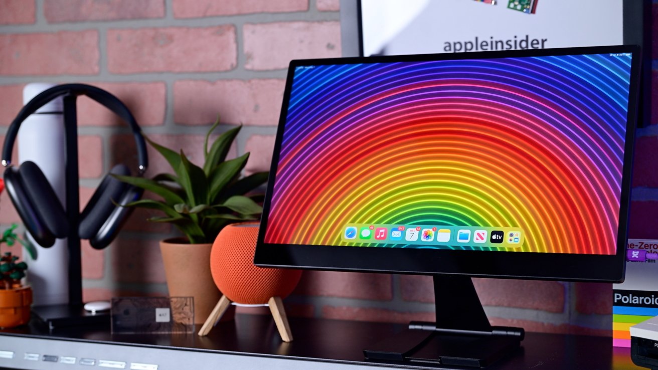 Computer monitor with a vibrant rainbow spiral design on screen, surrounded by headphones, a green plant, and an orange speaker on a desk with a brick wall background.