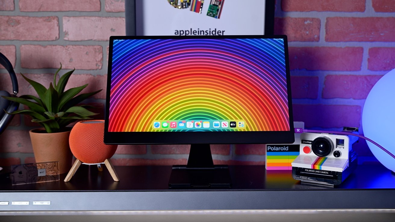 A computer monitor with a colorful rainbow pattern, a plant, an orange speaker, a Polaroid camera, and a glowing orb on a black desk against a brick wall backdrop.