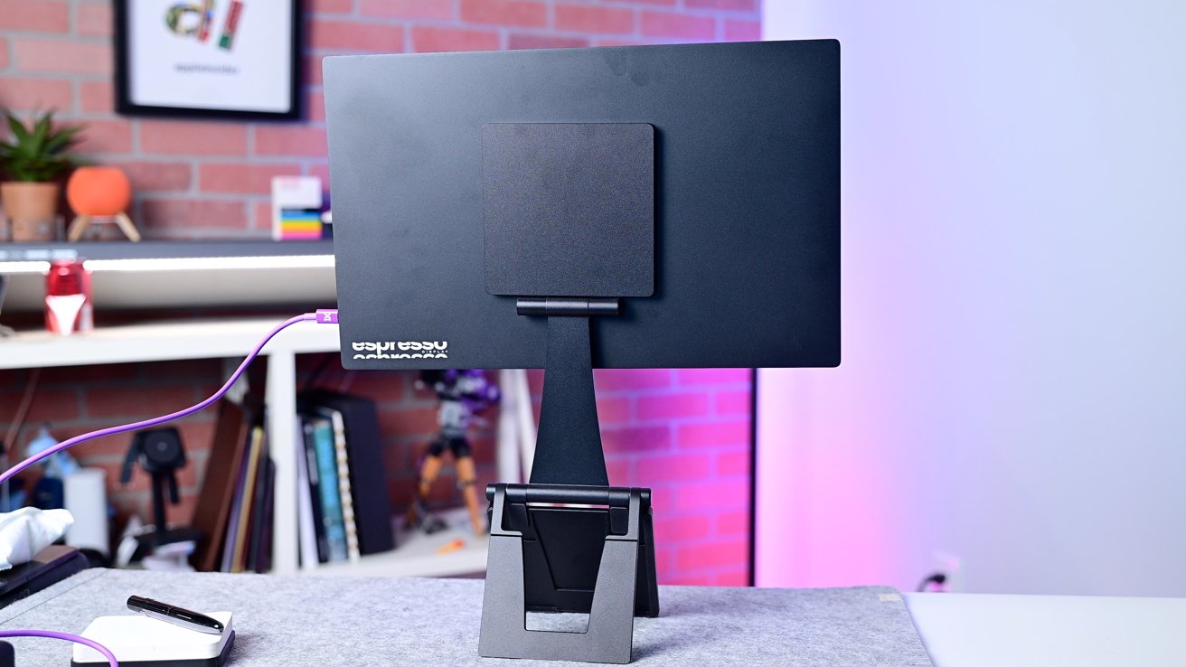 Rear view of a black monitor on a stand with a purple cable, in front of a brick wall with books and decor items.