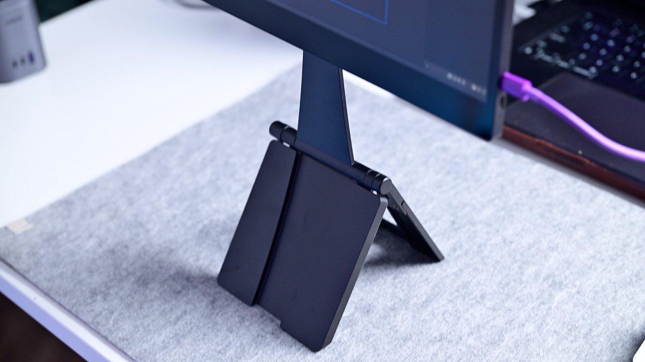 Monitor on a stand with two folding panels, placed on a gray desk mat near a laptop with a purple cable.