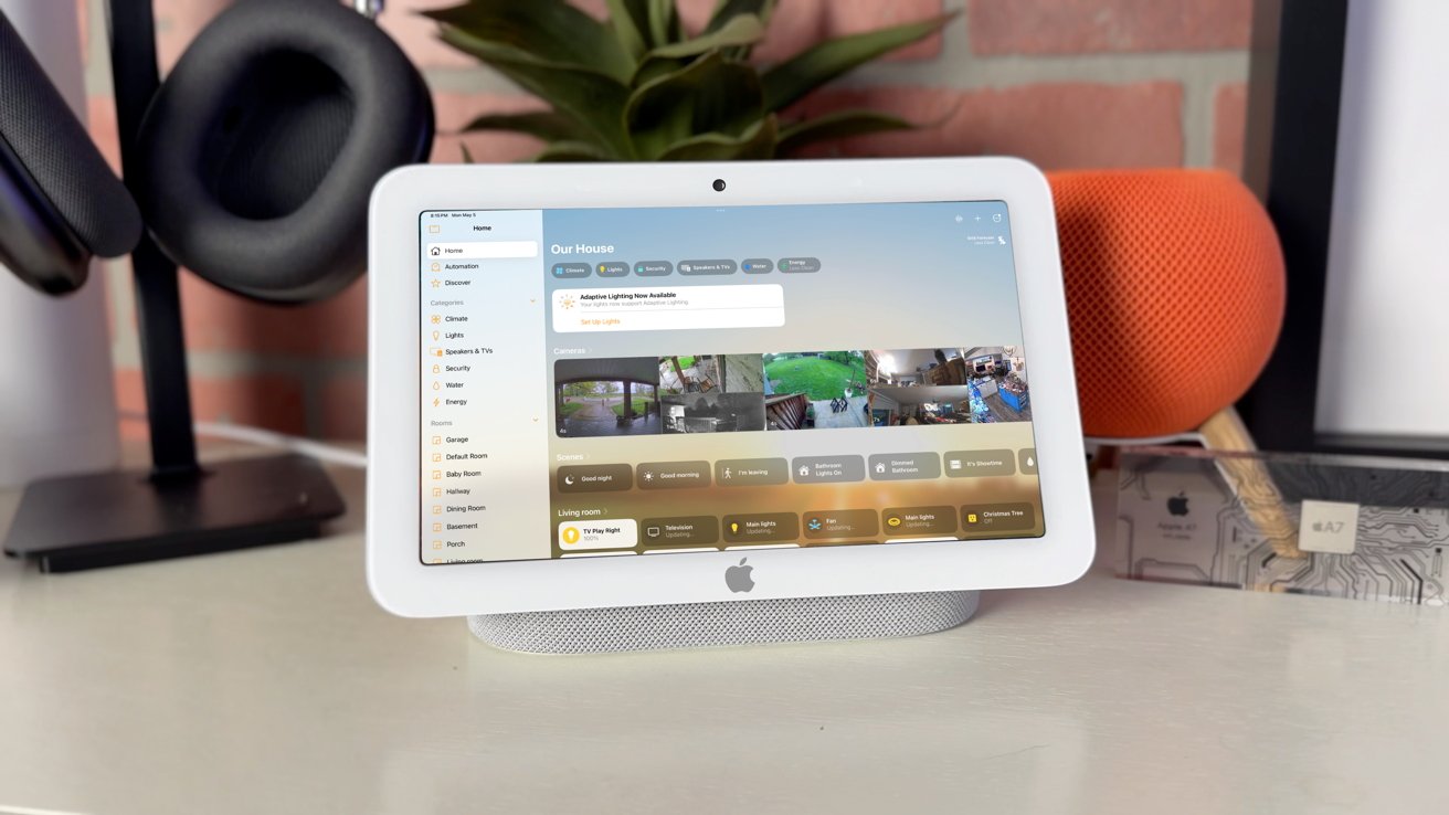 Tablet displaying home automation dashboard with security camera feeds and control options, placed on a desk. Surrounding items include headphones, an orange speaker, and a plant.