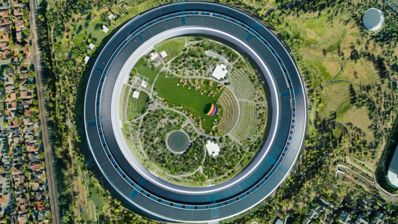 Aerial view of a circular building with a green courtyard, trees, and a colorful dome in the center, surrounded by urban and forested areas.