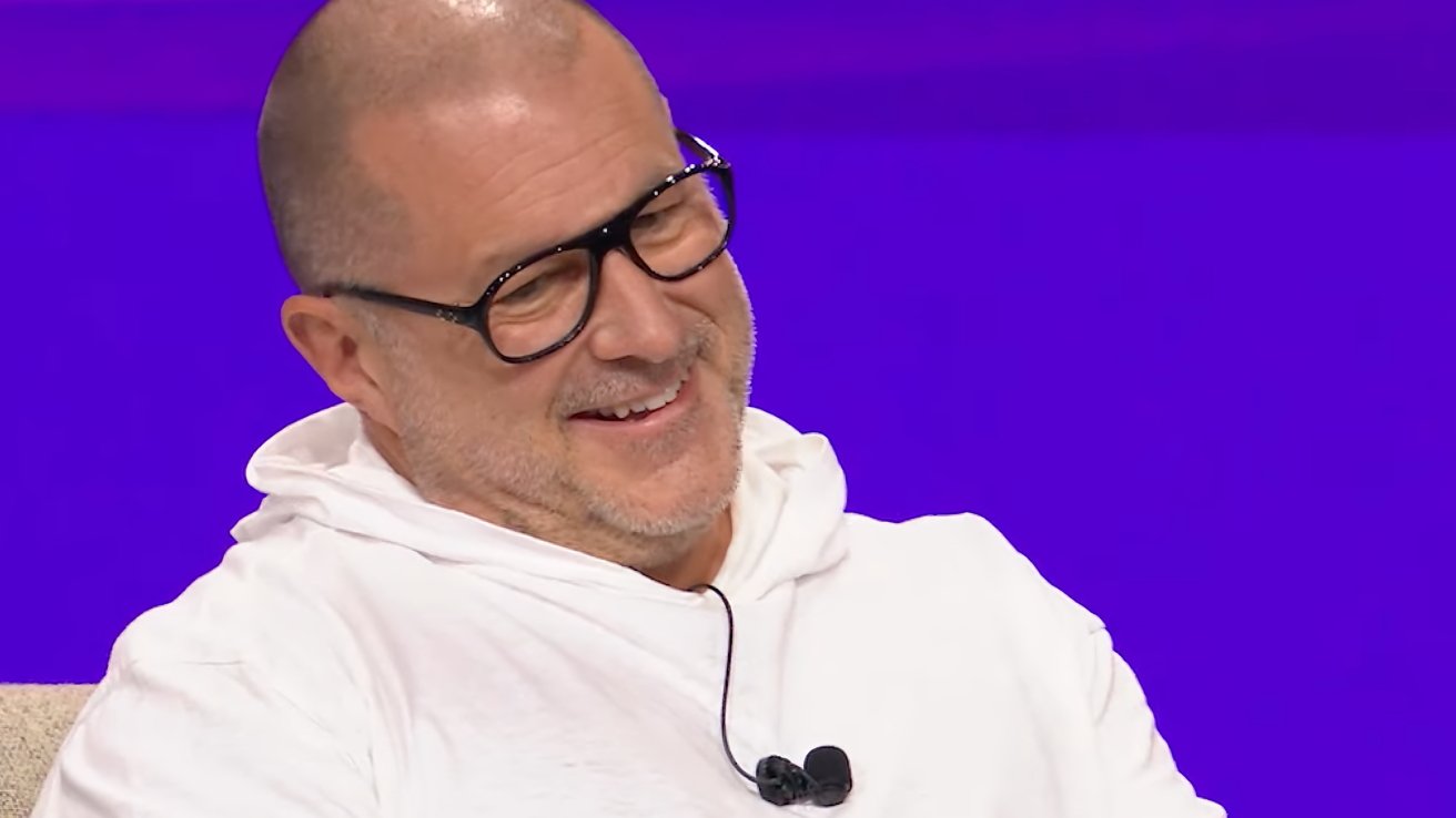 Smiling man in glasses and white hoodie, seated against a purple background, wearing a microphone.