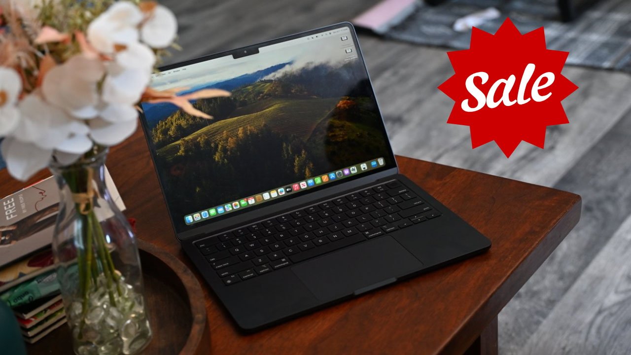 Open MacBook Air laptop on wooden table showing landscape wallpaper, vase with flowers nearby, red sale sticker in corner.