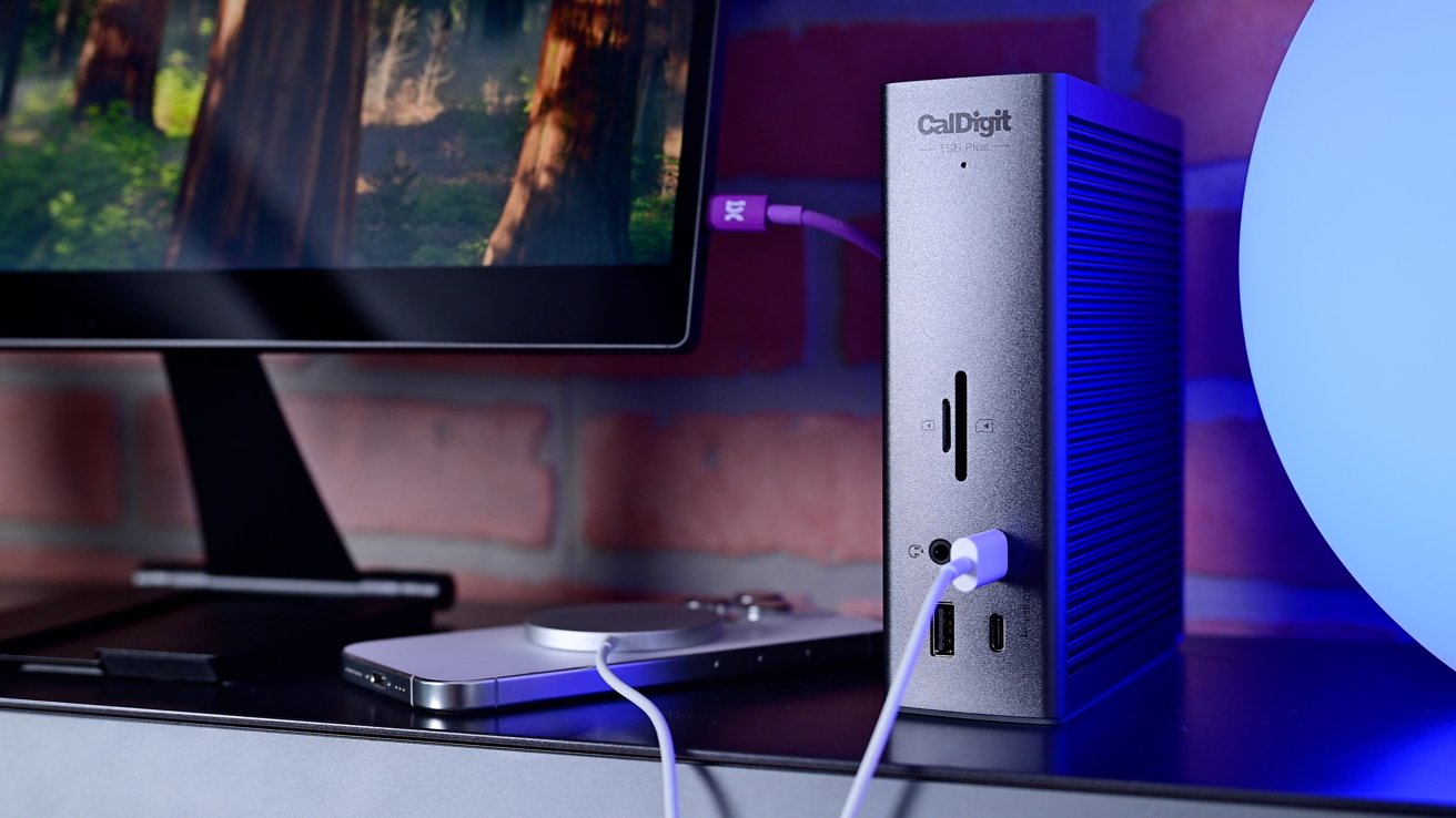 A vertical docking station with cables connected, next to a computer monitor displaying a forest scene, set against a brick wall illuminated by soft blue light.