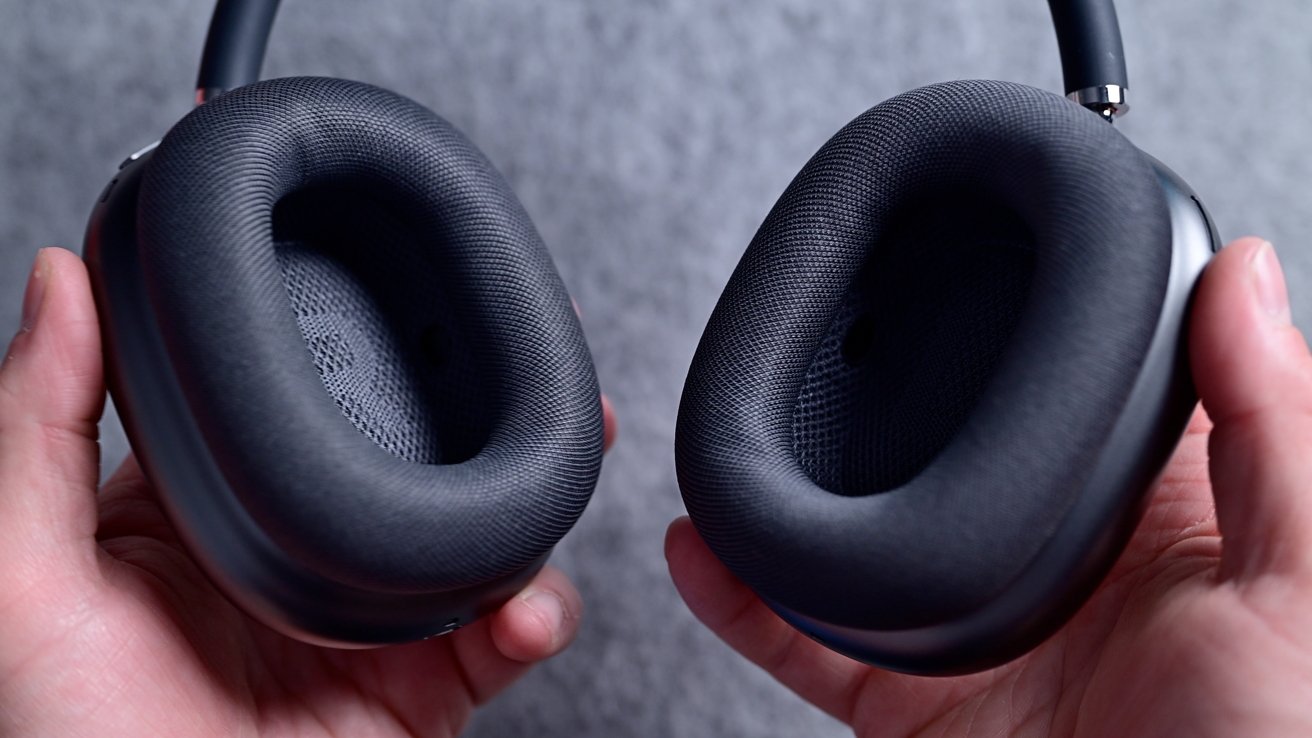 Two hands holding black over-ear headphones with soft, oval ear cushions against a gray textured background.