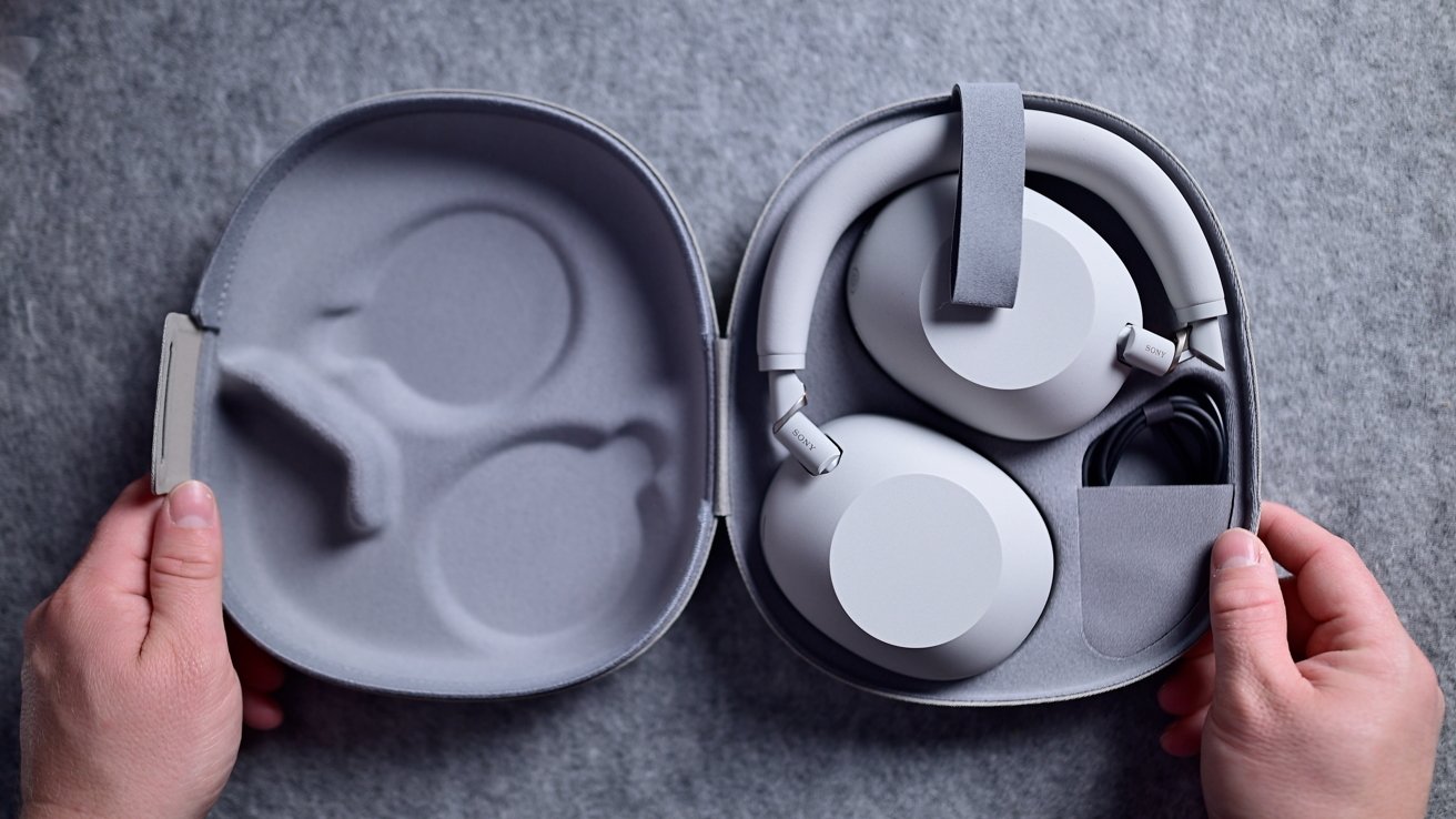 Hands holding an open gray case containing folded over-ear headphones and a neatly coiled cable against a gray fabric background.