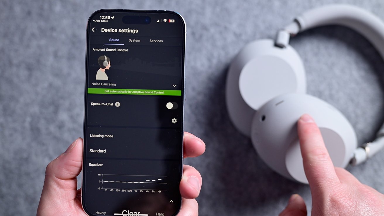 A hand holding a smartphone displaying device sound settings, including noise canceling. Another hand points at white over-ear headphones placed on a gray surface.