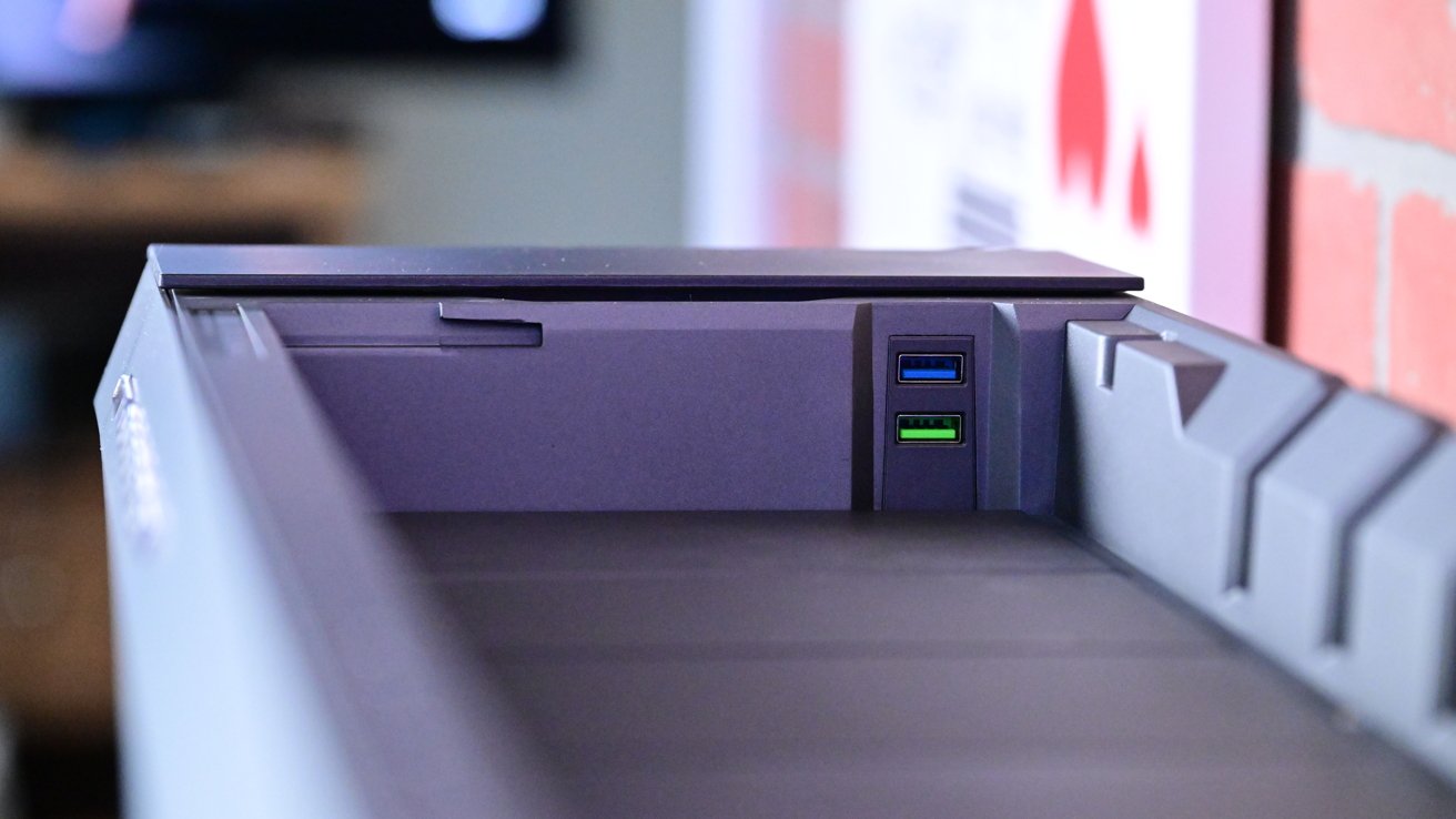 Gray computer tower with two USB ports, blue and green, on a blurred background featuring a brick wall and other objects.