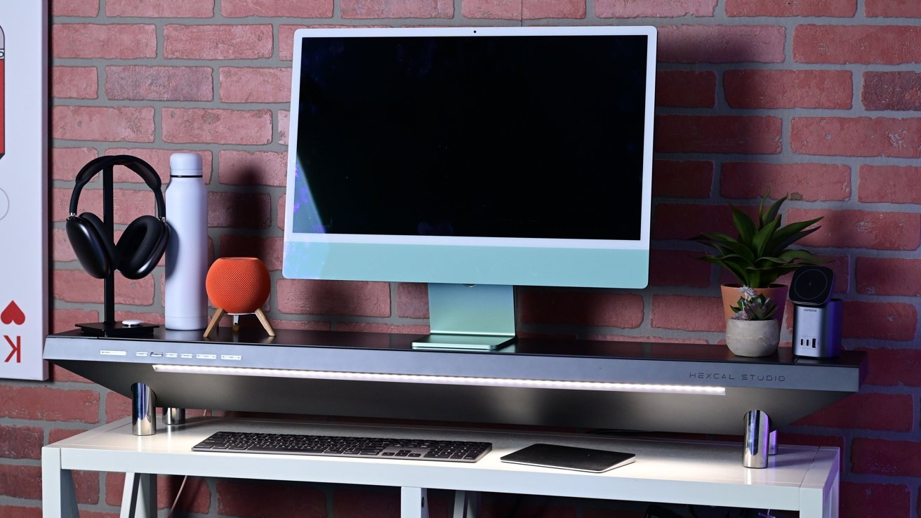 Computer monitor on a desk with keyboard, headphones, smart speaker, potted plant, and charger against a brick wall background.