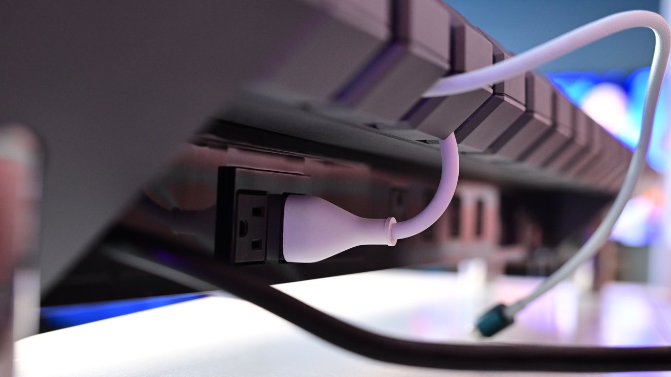 Close-up of a keyboard, with a white USB cable plugged into a nearby outlet, background softly blurred in colorful tones.