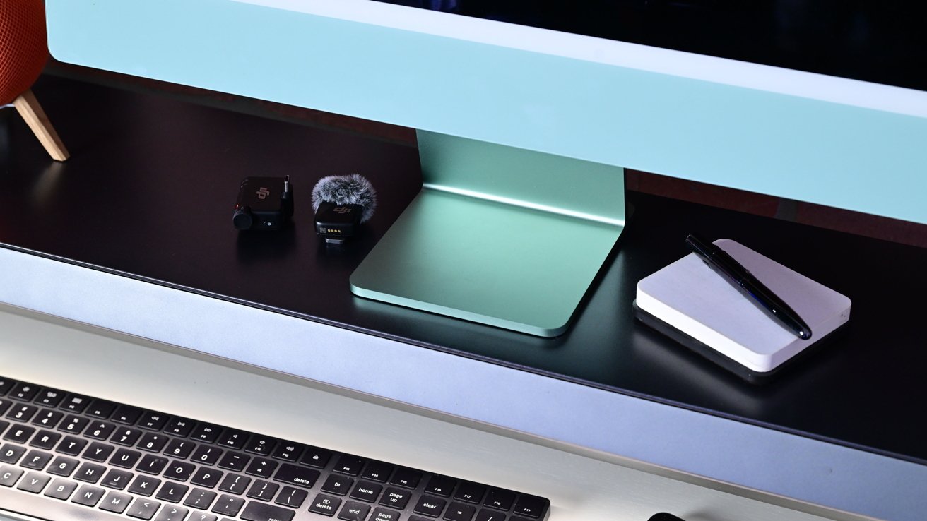 Desk setup with an iMac, wireless microphone, mini-laptop, and pen on a notebook.
