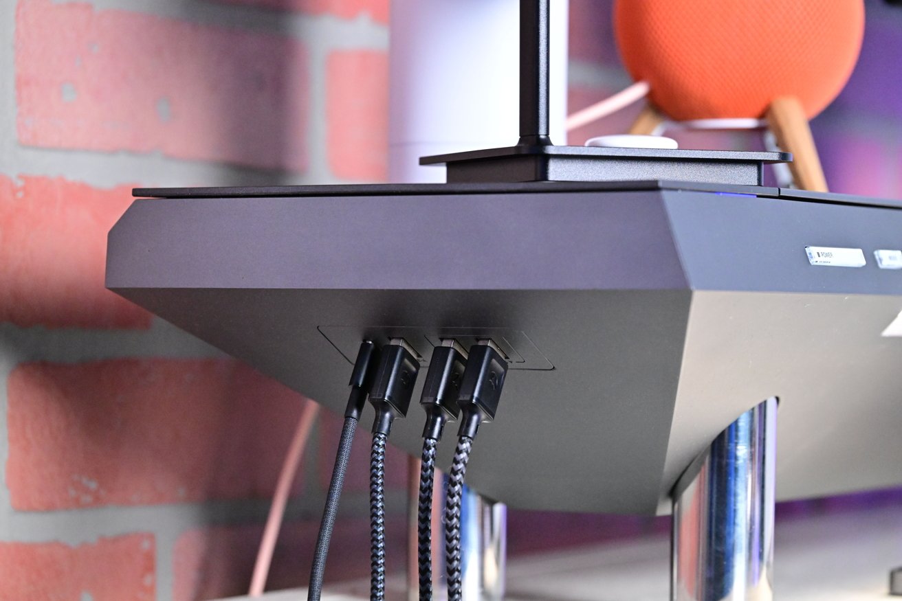 Black electronic device with multiple braided cables plugged in, set on a silver stand, with a brick wall background and a spherical orange object nearby.