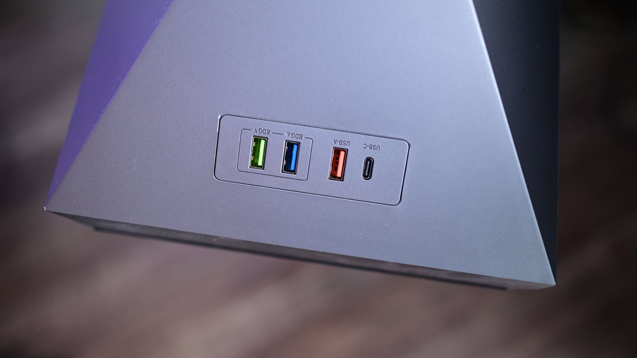 Triangular gray device with four USB ports: green, blue, red Type-A, and black Type-C, on a flat surface.