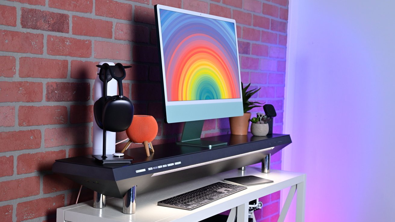 Desktop setup with a colorful rainbow screen, headphones stand, smart speaker, potted plants on a desk against a brick wall.