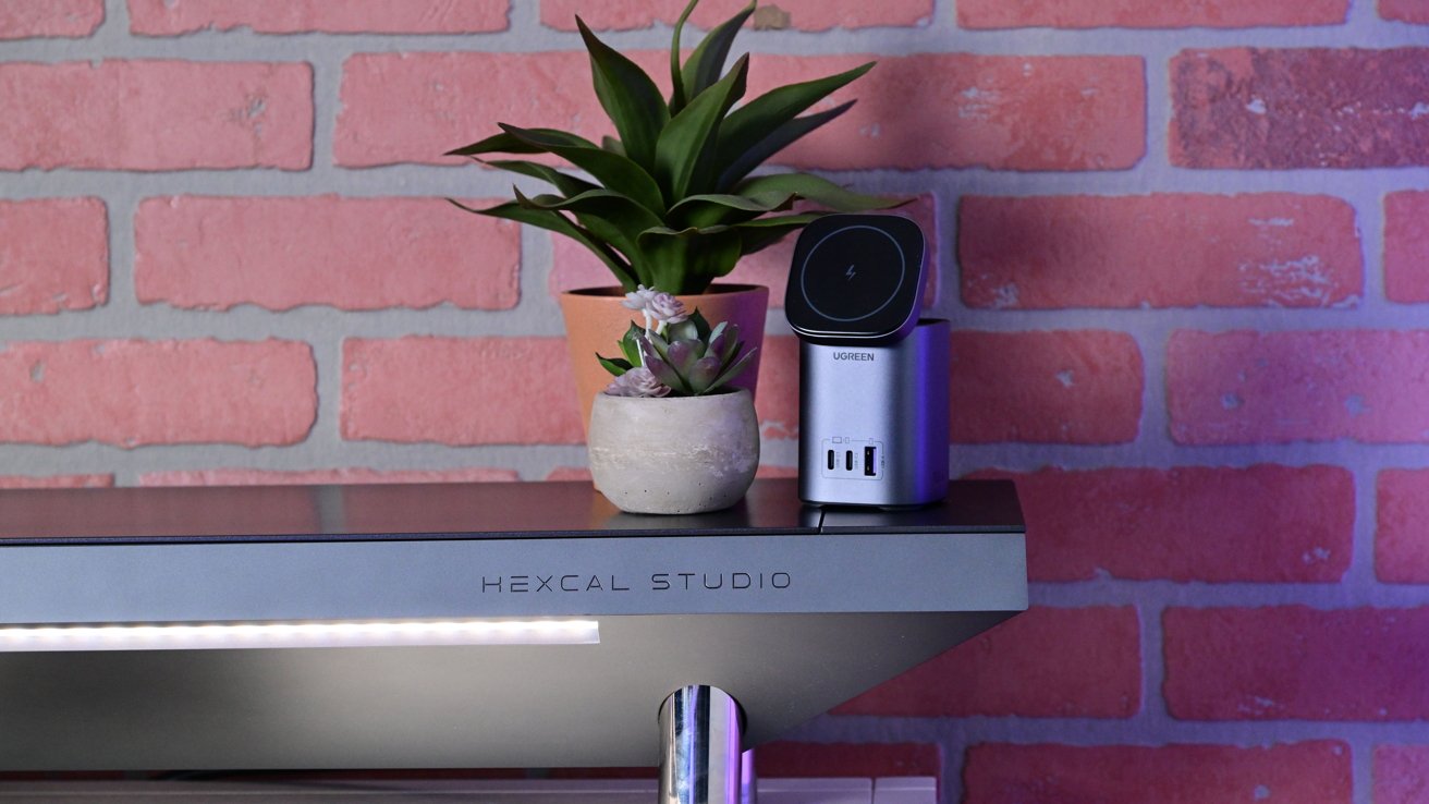 Desk with two potted plants and a gray charging station against a pink brick wall.