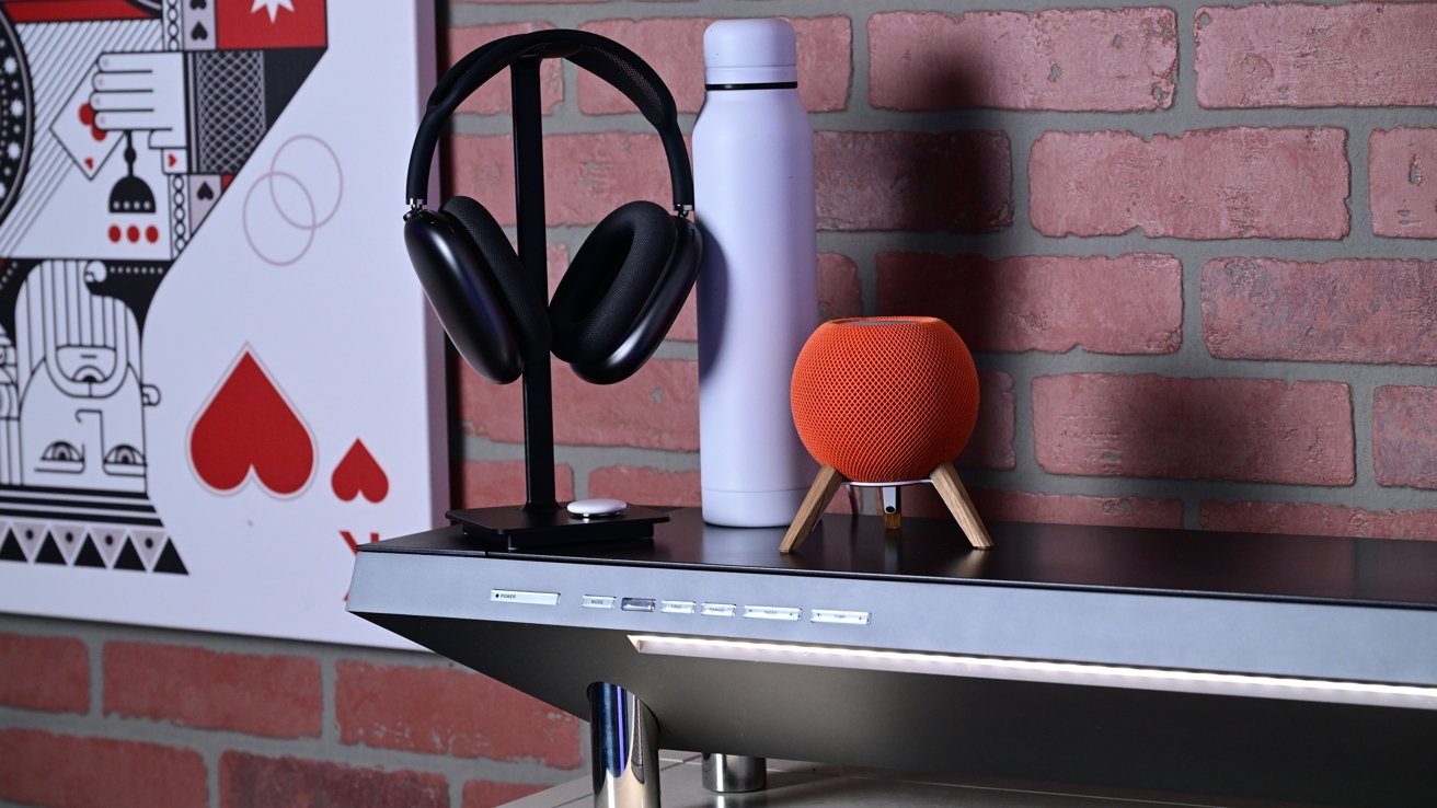 Headphones on stand, white bottle, orange speaker with wooden legs on black shelf, brick wall, and abstract art in background.