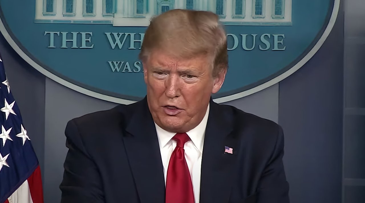 A man in a suit speaks at a podium in front of a blue background with 'The White House' written on it, American flag partially visible.