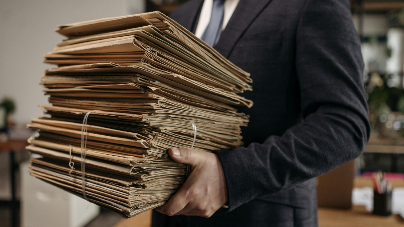 Person in a dark suit holding a large stack of tied, brown folders in an office environment.