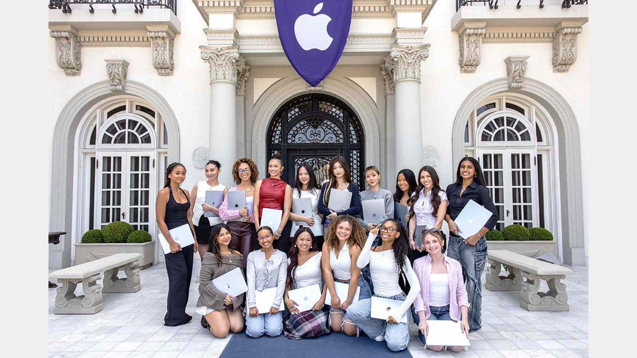 Group of young women holding laptops in front of a grand building with decorative pillars and a large logo banner.