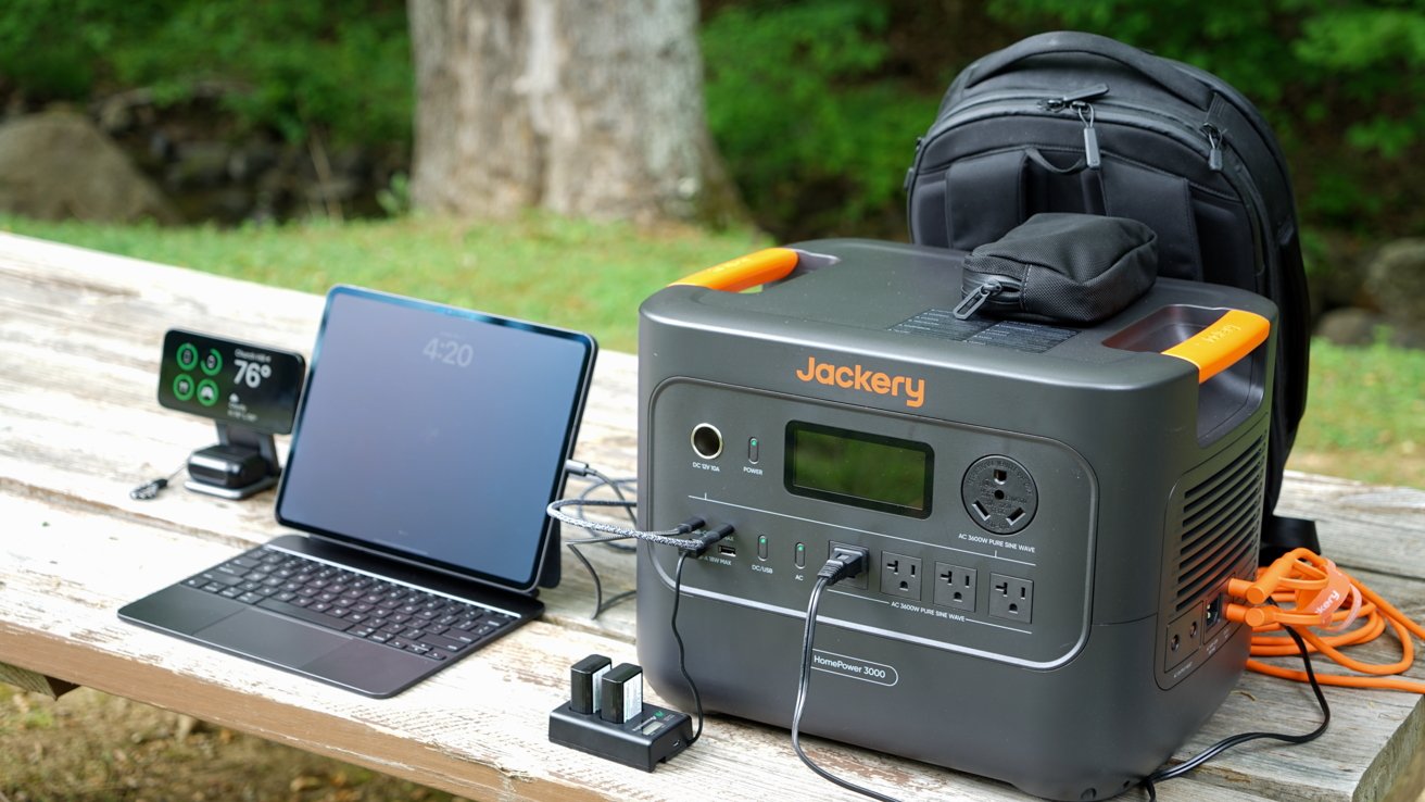 Portable power station with laptop, smartphone, and camera batteries charging on a wooden table outdoors; a backpack is placed behind the power station.