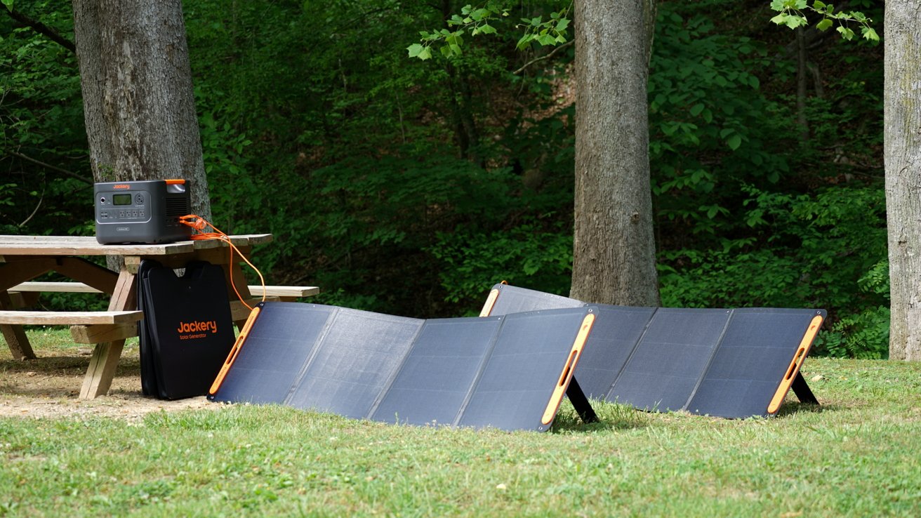 Portable generator connected to four solar panels on grass by a picnic table in a lush forest setting.