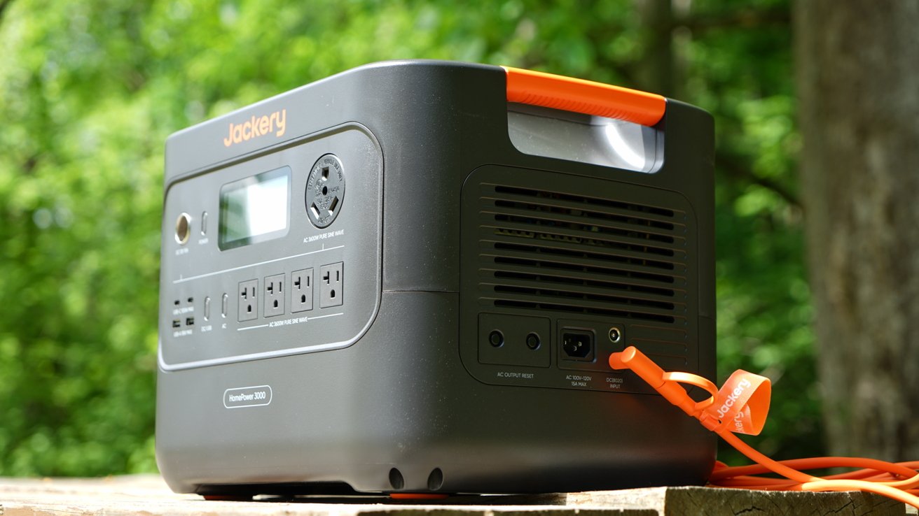 Portable power station with various outlets and an orange handle, placed outdoors on a wooden surface, surrounded by lush green foliage in the background.