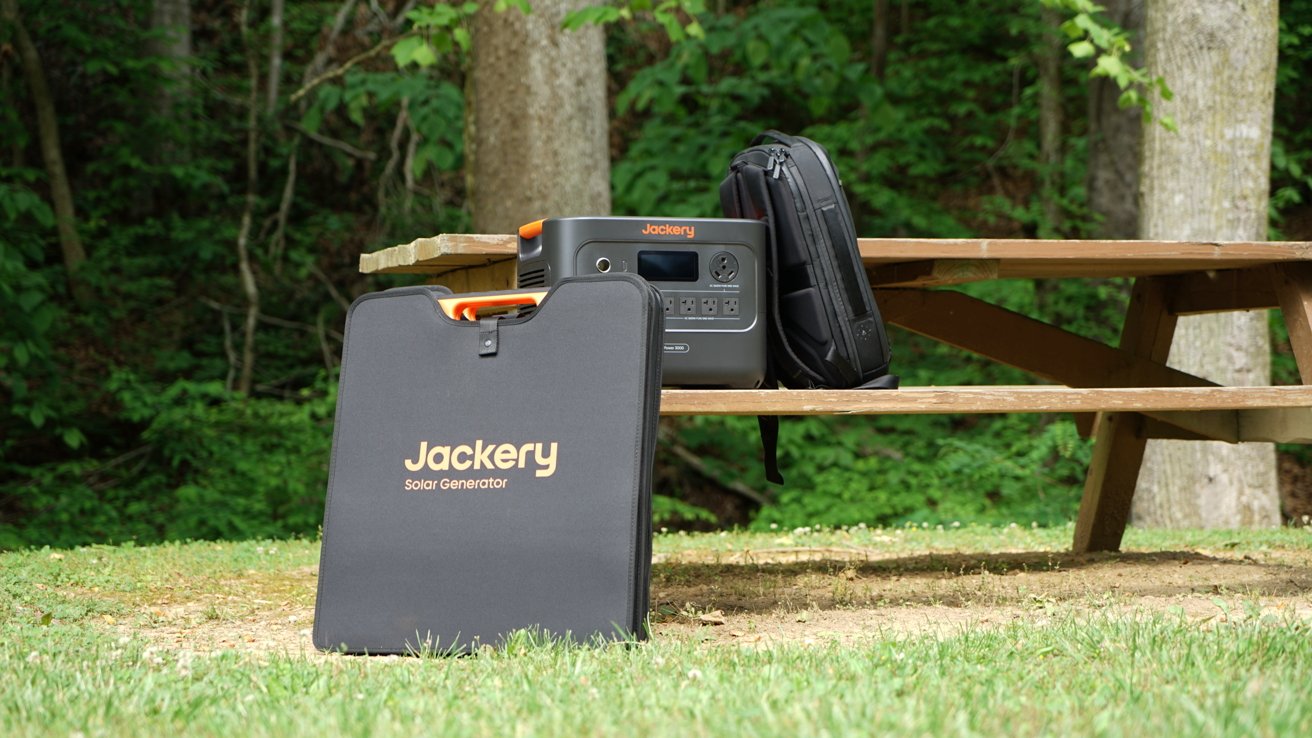 Portable solar generator, backpack, and folded solar panel on a wooden picnic table in a forested area with trees in the background.
