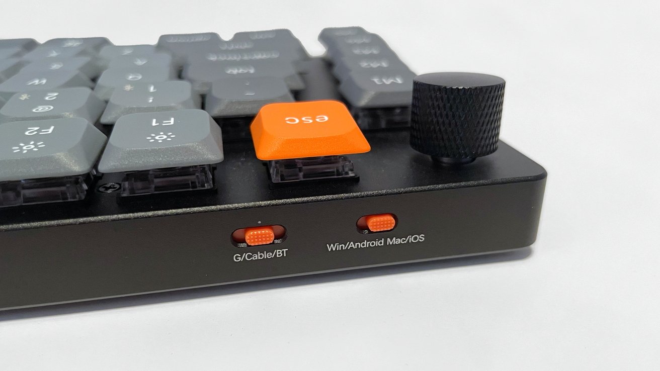 Gray mechanical keyboard keys with an orange escape key and a black knob. Two orange switches labeled for connectivity and operating system options are on the side.