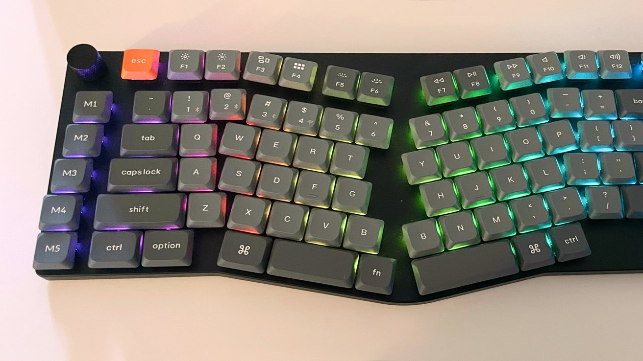 Ergonomic keyboard with split design, gray keys and colorful backlighting. The escape key is orange, and there's a black knob on the top left corner.
