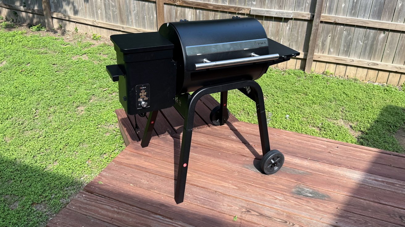 Black pellet grill on a wooden deck, surrounded by a grassy yard. A wooden fence is in the background.