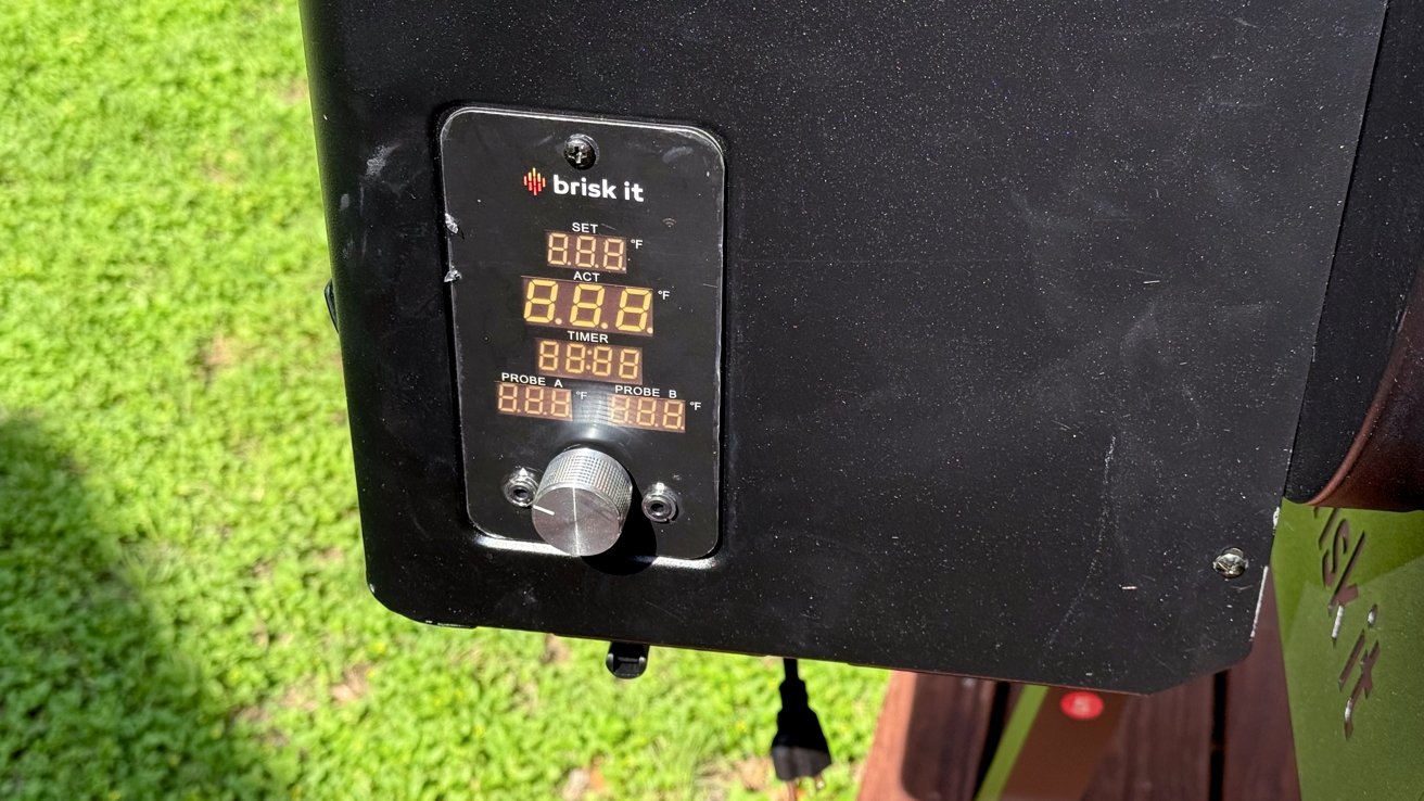 Black control panel with digital temperature display showing various settings and readings. Silver dial below displays adjustable controls. Green grass visible in the background.