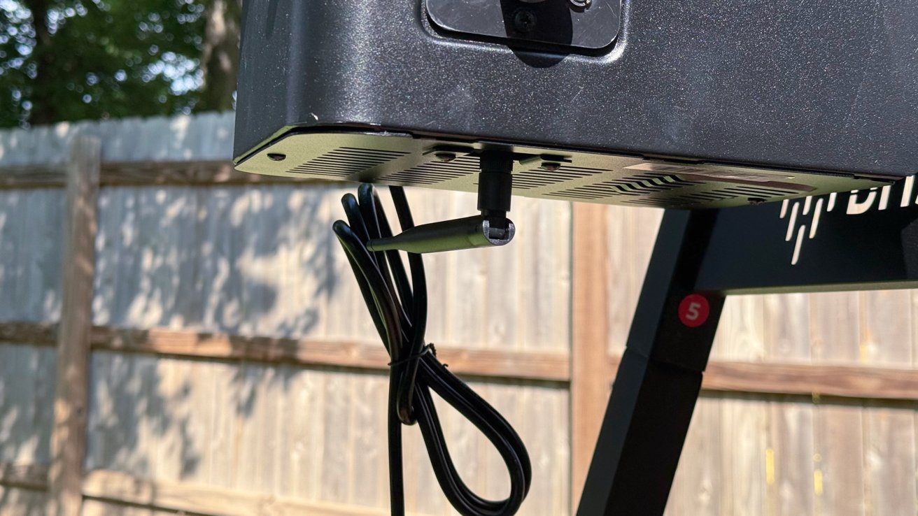 A large black speaker mounted outdoors with cables hanging underneath, set against a wooden fence with trees in the background.
