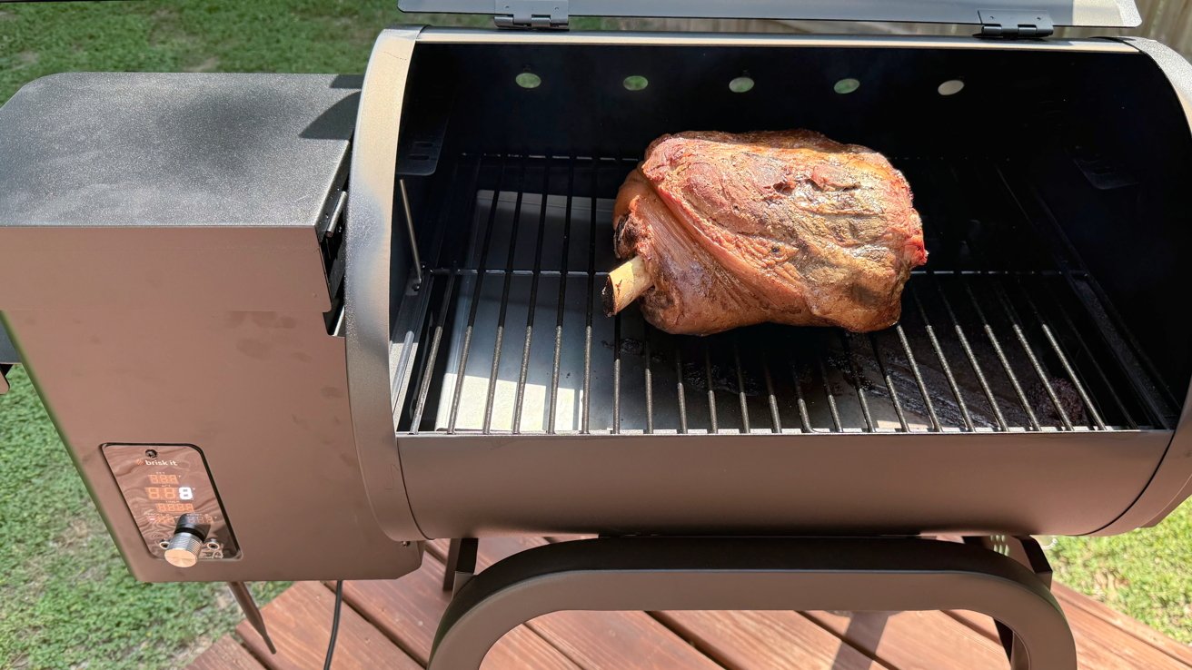 A large piece of meat on a grill inside a smoker, with digital control display, set on a wooden deck.