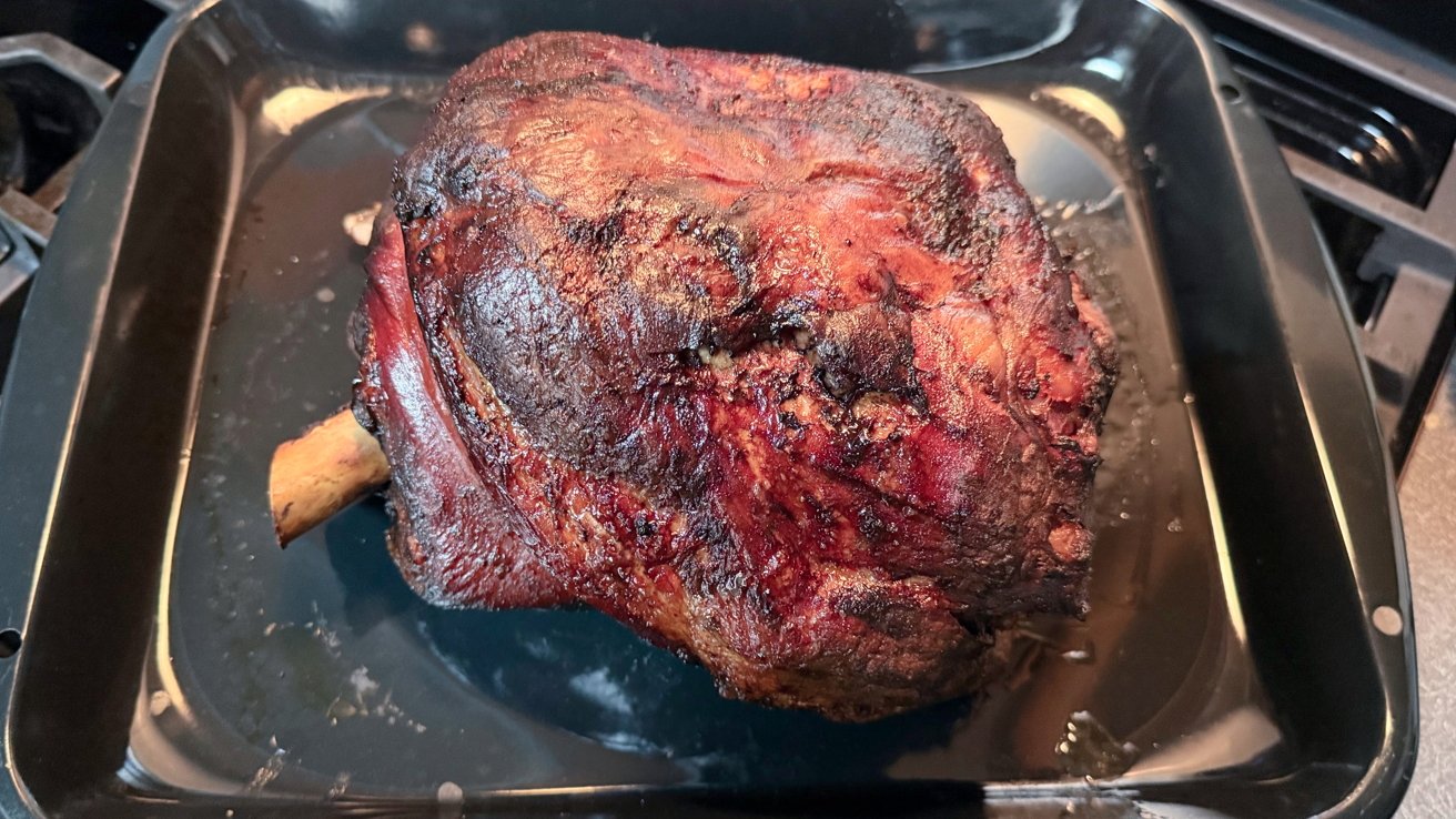Roasted, browned piece of meat with a visible bone end, resting in a black roasting pan with juices around it.