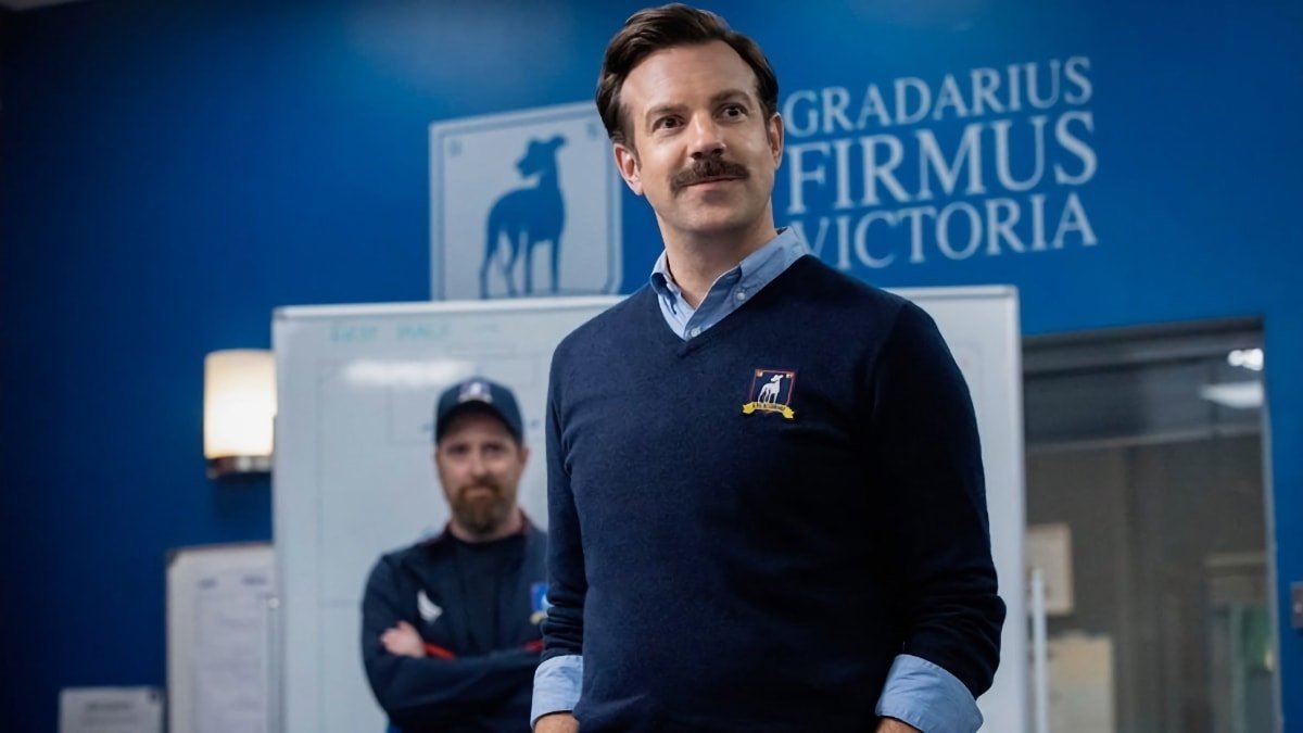 Smiling man with mustache in blue sweater stands in front of whiteboard and blue wall, another man in a cap is blurred behind him.