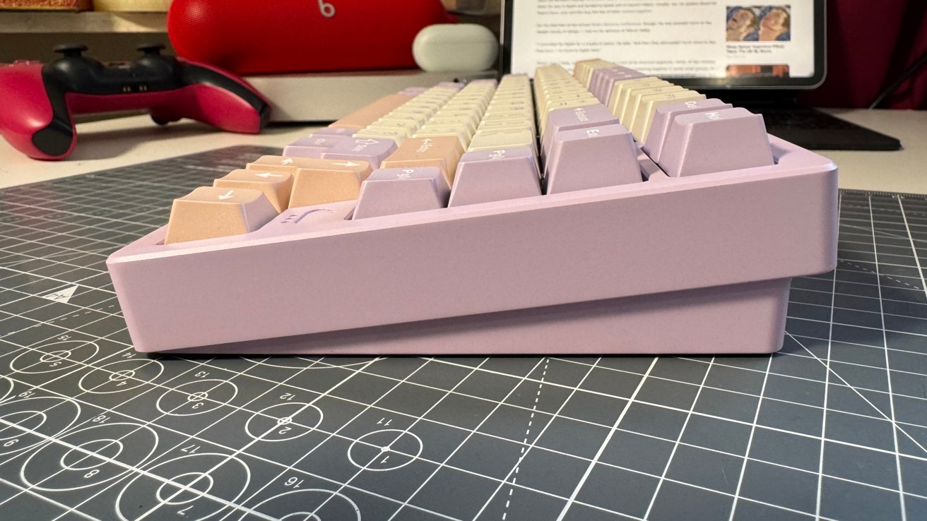 Side view of a pastel-colored mechanical keyboard on a grid-patterned surface, with a red and black gaming controller and a tablet in the background.