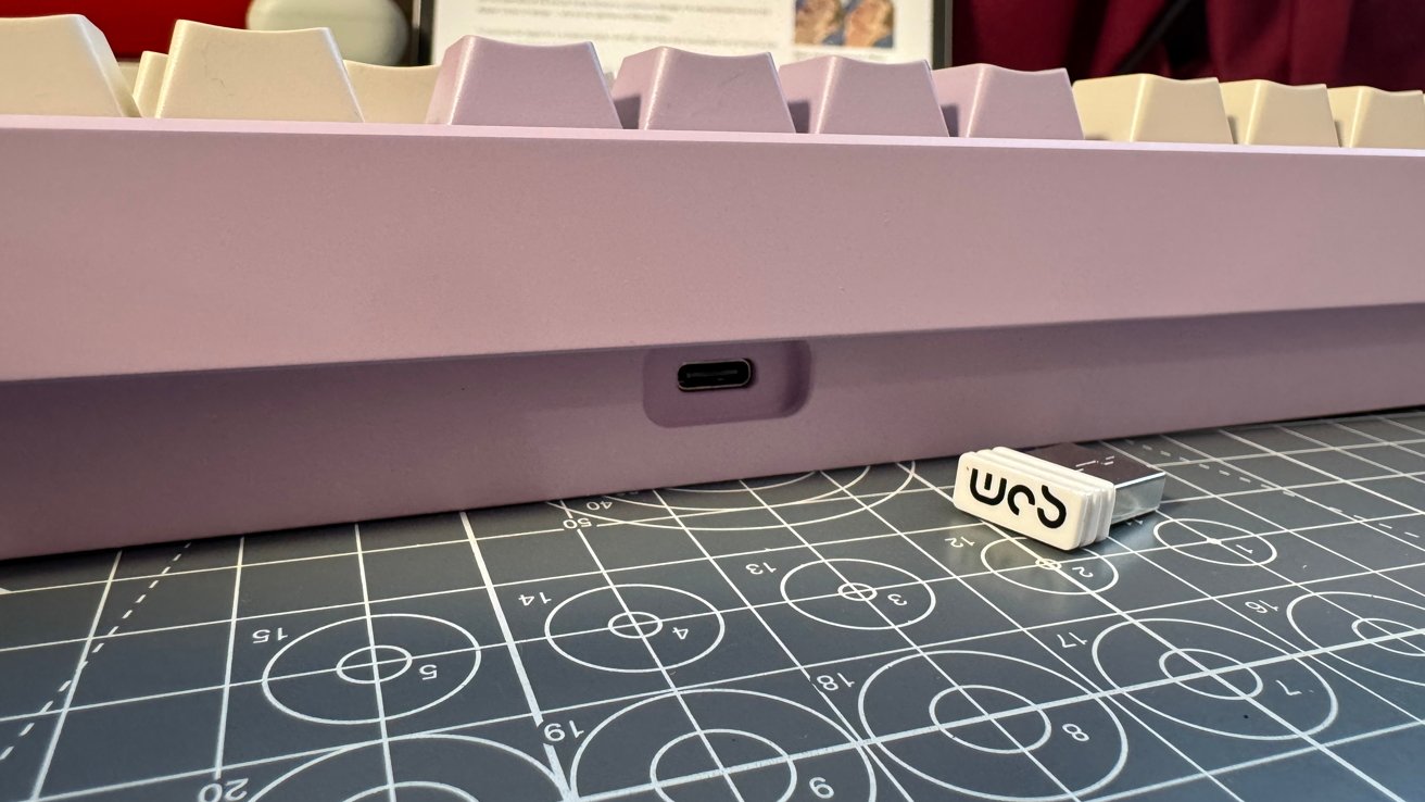 Purple mechanical keyboard with white keycaps, USB-C port visible. A USB drive rests on a blue cutting mat with grid and circle patterns.