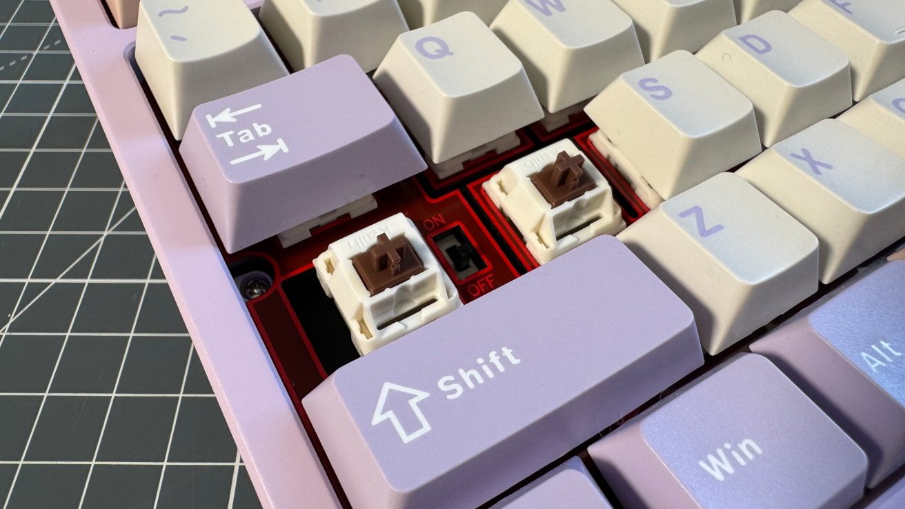 Close-up of a pastel-colored mechanical keyboard with some keys removed, exposing brown switches. A visible switch labeled ON/OFF is set to OFF, all on a grid-patterned surface.
