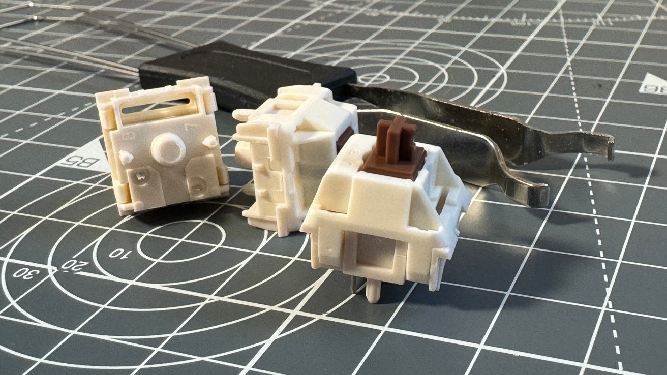 Three white mechanical keyboard switches, one with a brown stem, on a grid cutting mat with metal tools in the background.