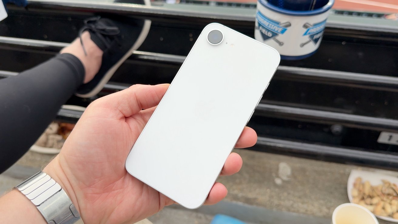 A hand holding a white smartphone with a single camera lens, sitting outdoors.