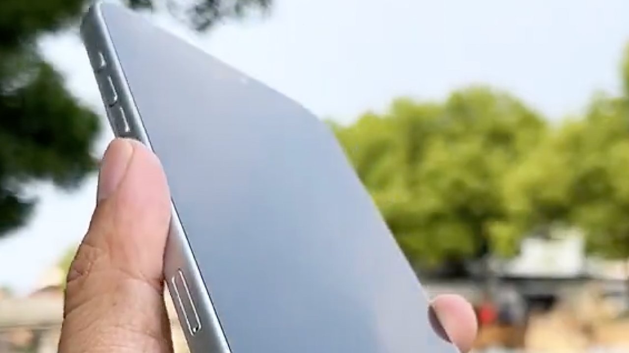 A hand holding a slim silver smartphone outdoors, with green trees blurred in the background.