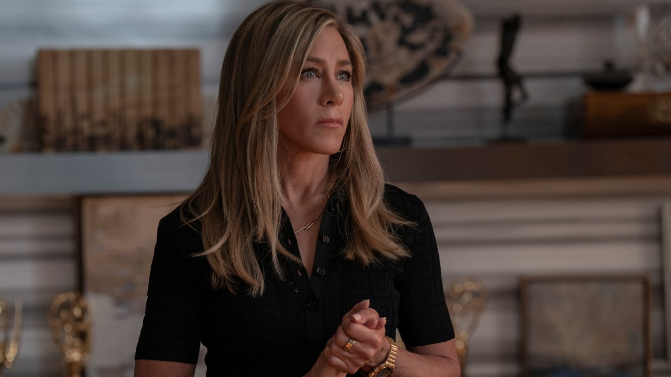 Person with long blonde hair wearing a black top stands indoors, looking thoughtfully to the side. Shelves with decorative items are in the background.